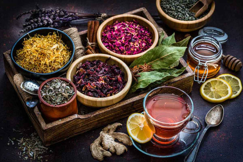 Bowls of dried herbs, a glass of tea, and lemon slices on a dark surface.