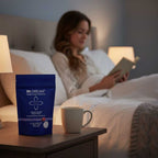 Woman reading a book in bed with a cup and IN DREAM pouch on a nightstand.