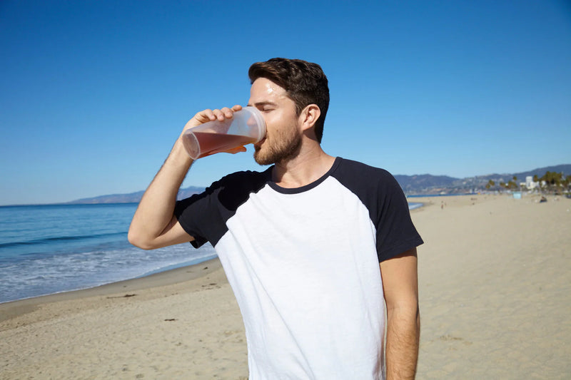 Man drinking from a cup on a beach with clear blue sky and ocean.